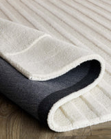 Close-up of a rolled-up, plush, off-white area rug with a contrasting black border, displaying its texture and thickness, set against a wooden floor background.