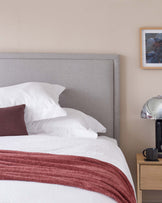 A modern bedroom featuring a large upholstered headboard in grey fabric, flanked by a minimalistic wooden bedside table with an open shelf and a single drawer. The bedding is accented with a plush burgundy throw running across a plain white duvet cover. A sleek, metallic table lamp with a round shade adds a contemporary touch to the setting.