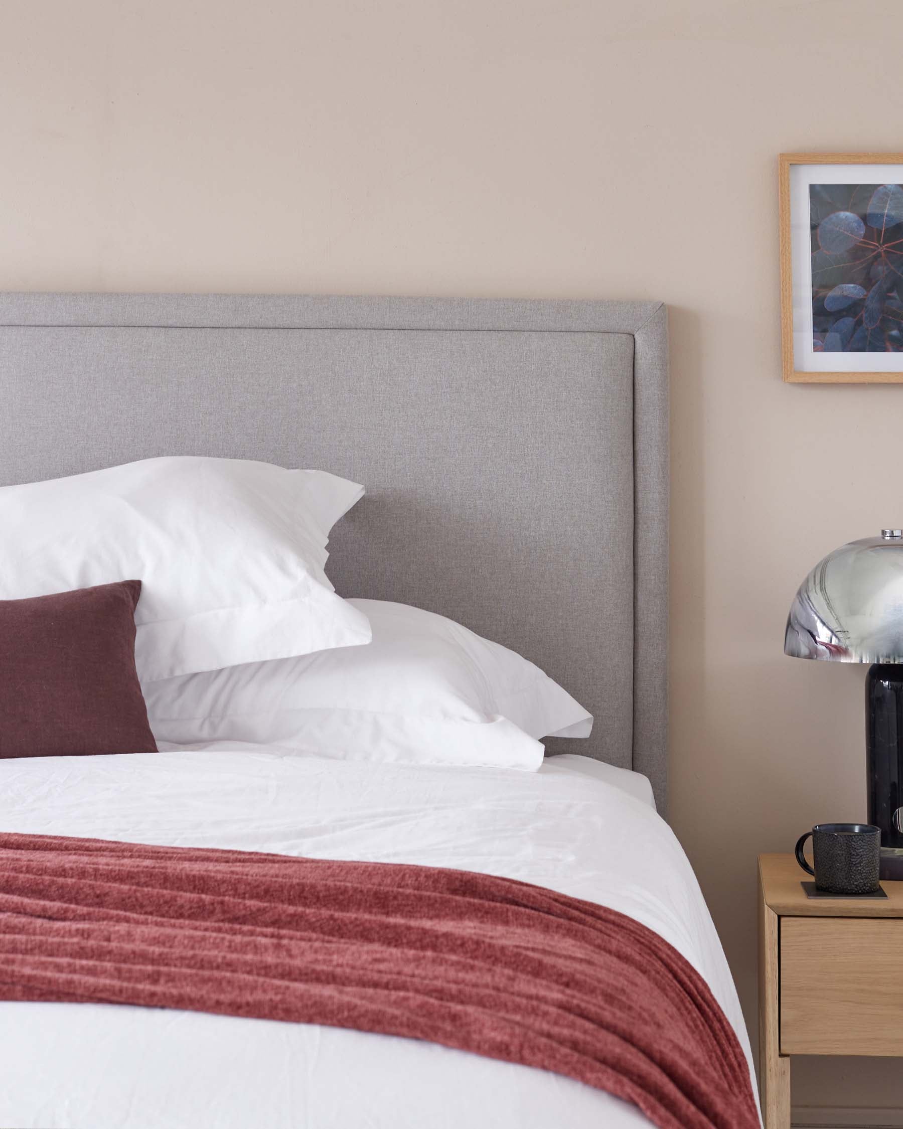 A modern bedroom with a large bed featuring a grey upholstered headboard. Beside the bed is a light wood nightstand with a simple, clean design. A metallic table lamp with a dome-shaped shade adds a touch of contemporary elegance.