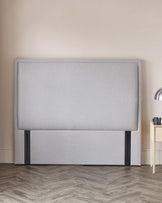 Contemporary grey upholstered headboard with a minimalist design and black metal legs, accompanied by a light wooden bedside table with a black vase.