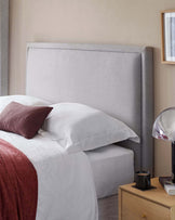 A modern bedroom featuring a large upholstered headboard in a neutral grey fabric. A wooden bedside table with a simple drawer and a metal handle is visible beside the bed, matching the understated and contemporary style of the room.