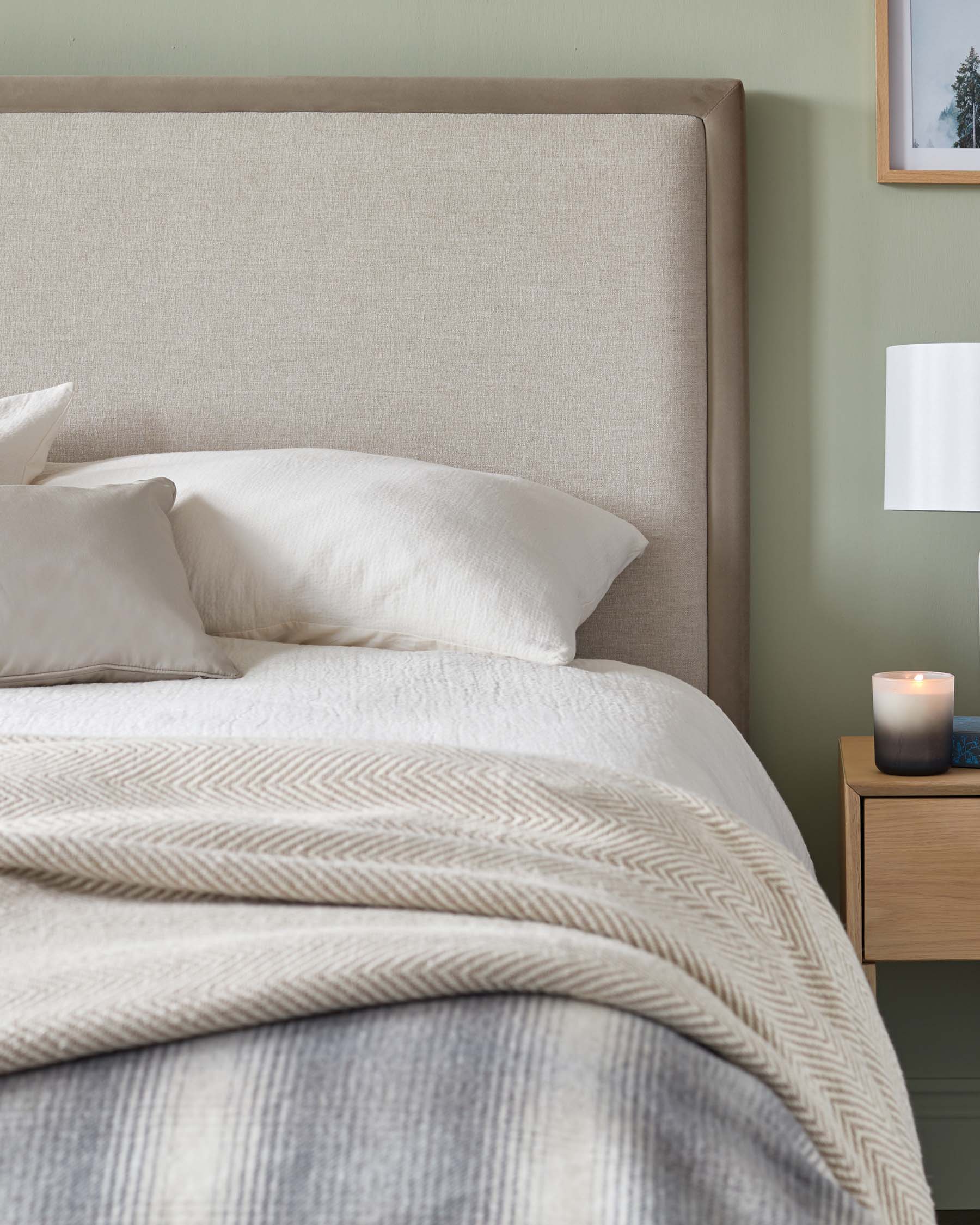 A contemporary beige upholstered headboard paired with a matching neutral-toned bed set. To the side is a minimalist natural wood bedside table featuring a single drawer, complemented by a white lamp with a cylindrical shade, and a small lit candle atop. The scene is accented with a framed artwork on the wall, contributing to a serene and sophisticated bedroom ambiance.