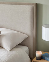 "Elegant upholstered headboard in a neutral beige fabric, paired with a matching bed dressed in white linens. A wooden bedside table holds a candle, a book, and a simple white lampshade."