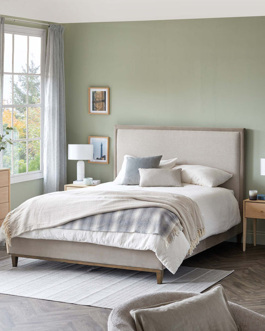 Upholstered queen-sized bed with a high headboard, two-tone neutral bedding, and a fringed throw. A matching wooden nightstand with a simple table lamp and two drawers. There's also a plush area rug beneath the bed.