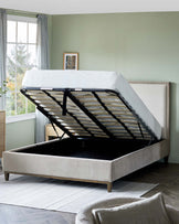 Elegant modern upholstered bed with a lift-up storage system. The bed features a light grey fabric finish, a tufted headboard, and slim wooden legs. The mattress is raised to reveal a spacious under-bed storage compartment with a black interior, supported by a sturdy metal frame with wooden slats. The bedroom setting has a muted colour scheme with a soft green wall, grey curtains, and a framed picture above the headboard, all complementing the contemporary aesthetic of the bed.