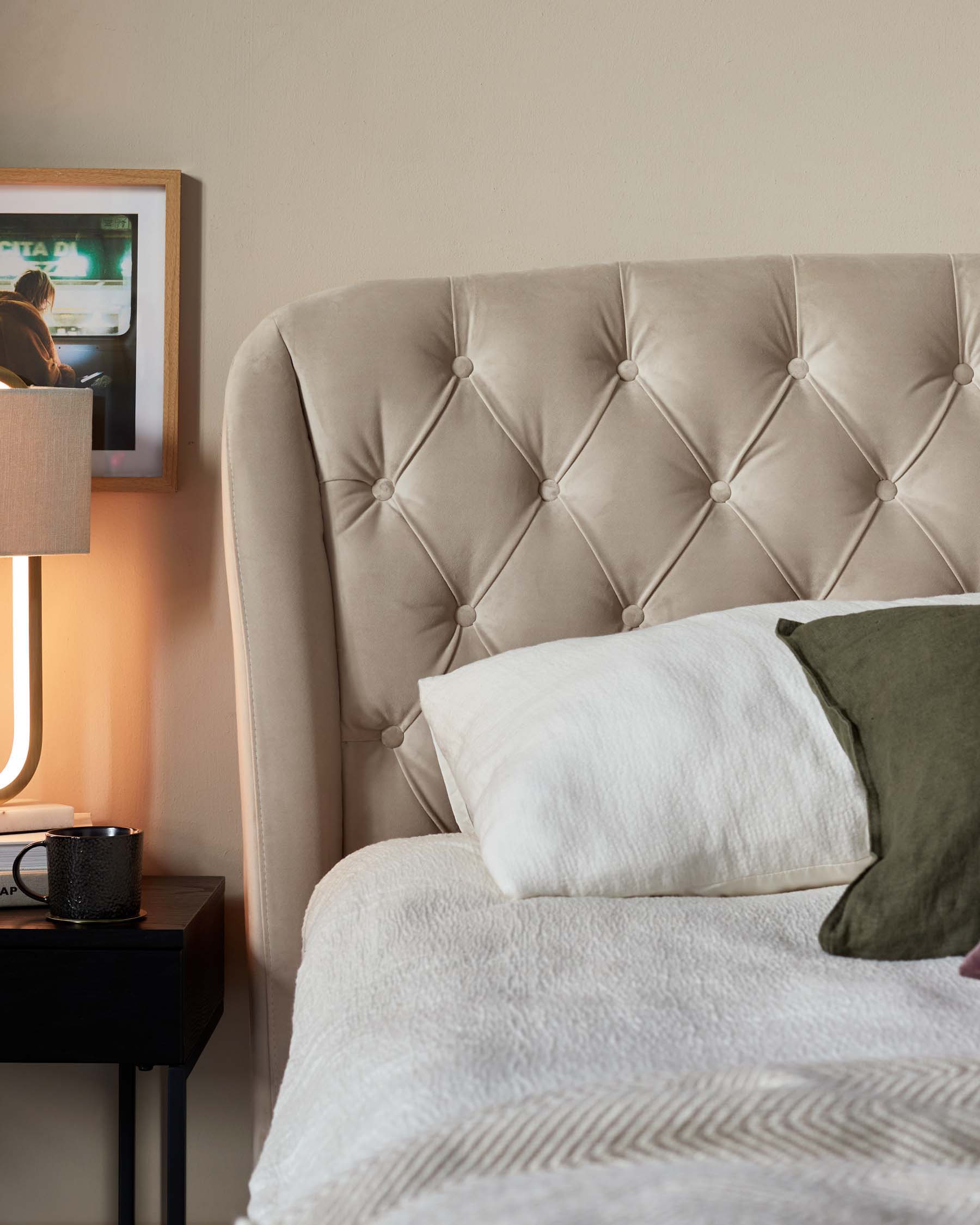 Elegant tufted headboard in a soft beige fabric, paired with a simple black nightstand featuring a contemporary lamp and decorative accessories, beside a bed adorned with textured white bedding and accent pillows.