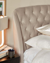 Elegant beige upholstered headboard with tufted button design, paired with a neutral-toned bedside table with a textured ceramic mug and stacked books, beside a cosy bed dressed in white linens.