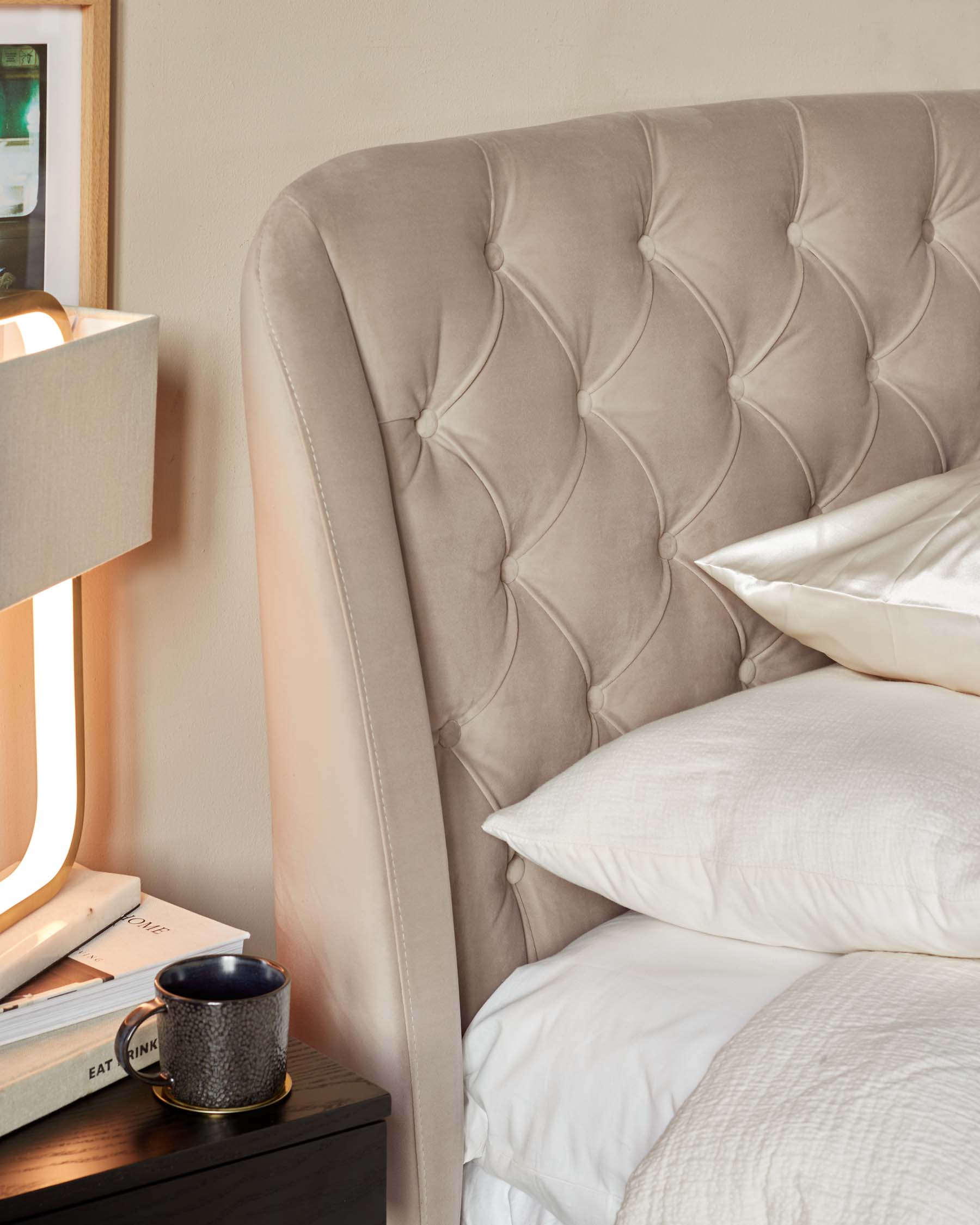 Elegant beige upholstered headboard with tufted button design, paired with a neutral-toned bedside table with a textured ceramic mug and stacked books, beside a cosy bed dressed in white linens.