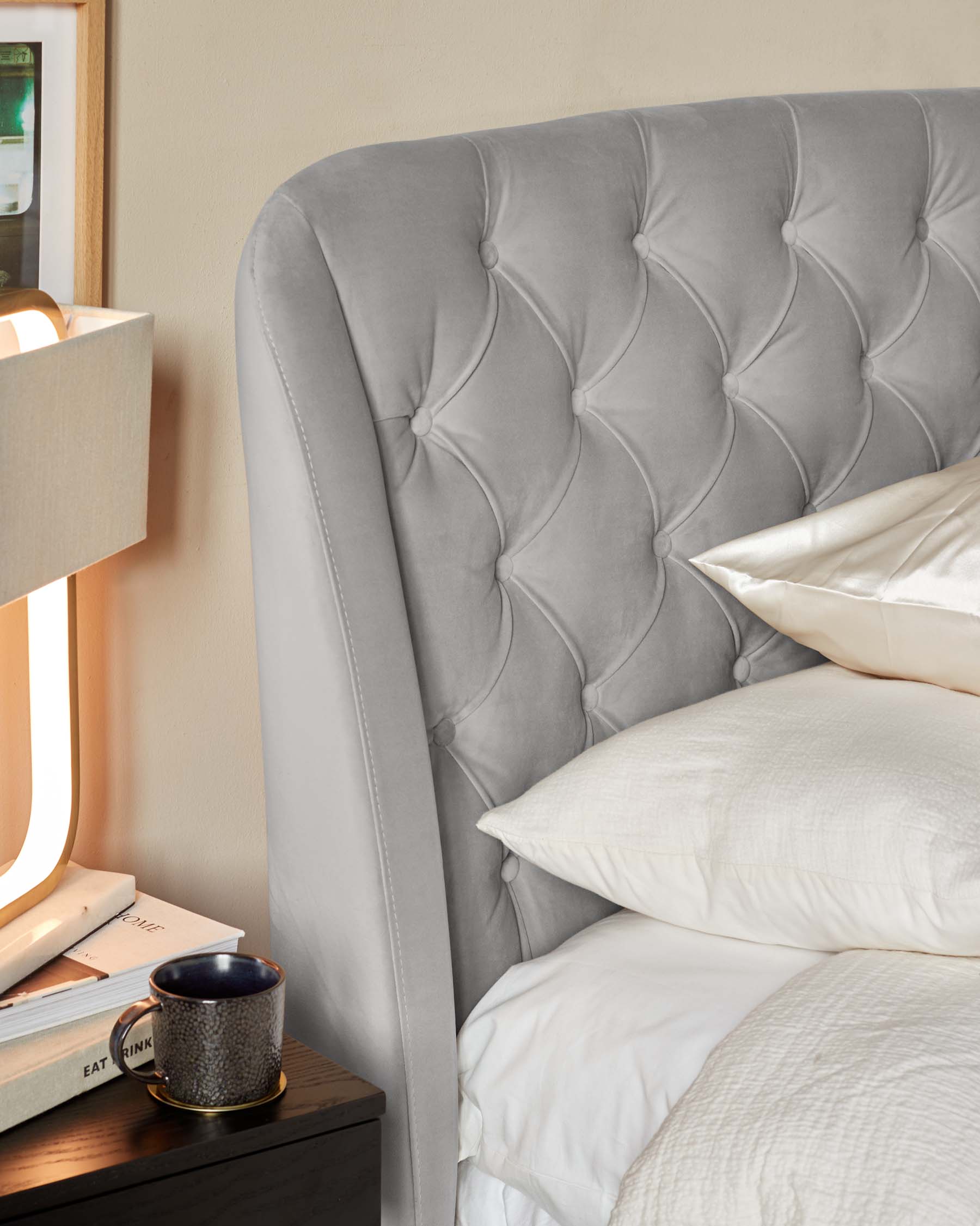 Elegant grey tufted headboard with curved wingback design, featuring button accents and visible stitching details, paired with a simplistic dark wood bedside table.