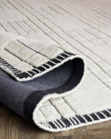 Close-up of a luxurious, textured area rug with a geometric pattern, in shades of cream and charcoal, partially rolled up on a wooden floor.