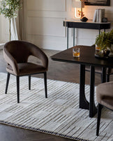 Elegant modern furniture set comprising a dark wooden dining table with a rectangular top and black metal legs, and a pair of luxurious upholstered armchairs with a smooth, curved design and black metal legs, all placed on a textured grey and white area rug with a geometric pattern. The scene is complemented by a sleek black console table in the background with a lamp, books, and decorative items, enhancing the sophisticated and contemporary ambiance.