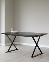 Modern dark wood dining table with stylish X-shaped metal legs, topped with plates and a glass on a minimalistic floor.