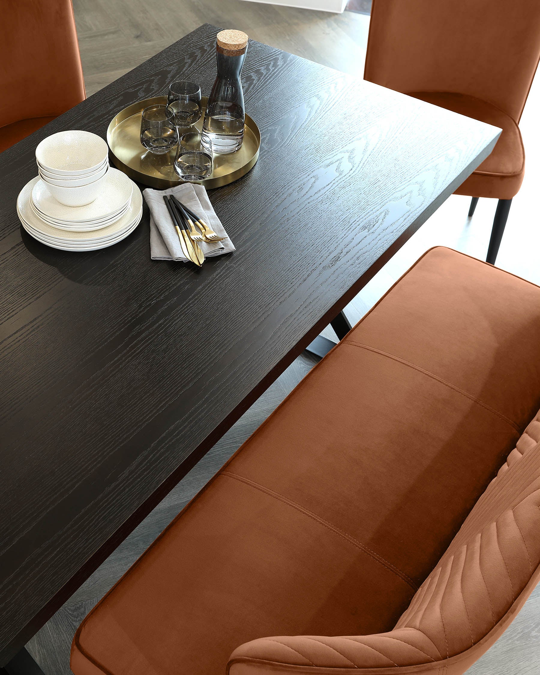 Modern dining room featuring a dark wood table with visible grain texture and two tan upholstered chairs with vertical channel stitching. A tray with glassware and dinnerware is arranged on the table.