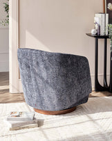 Modern curved-back accent chair in textured blue-grey fabric on a swivel wooden base, paired with a round black side table displaying books and a small potted plant.