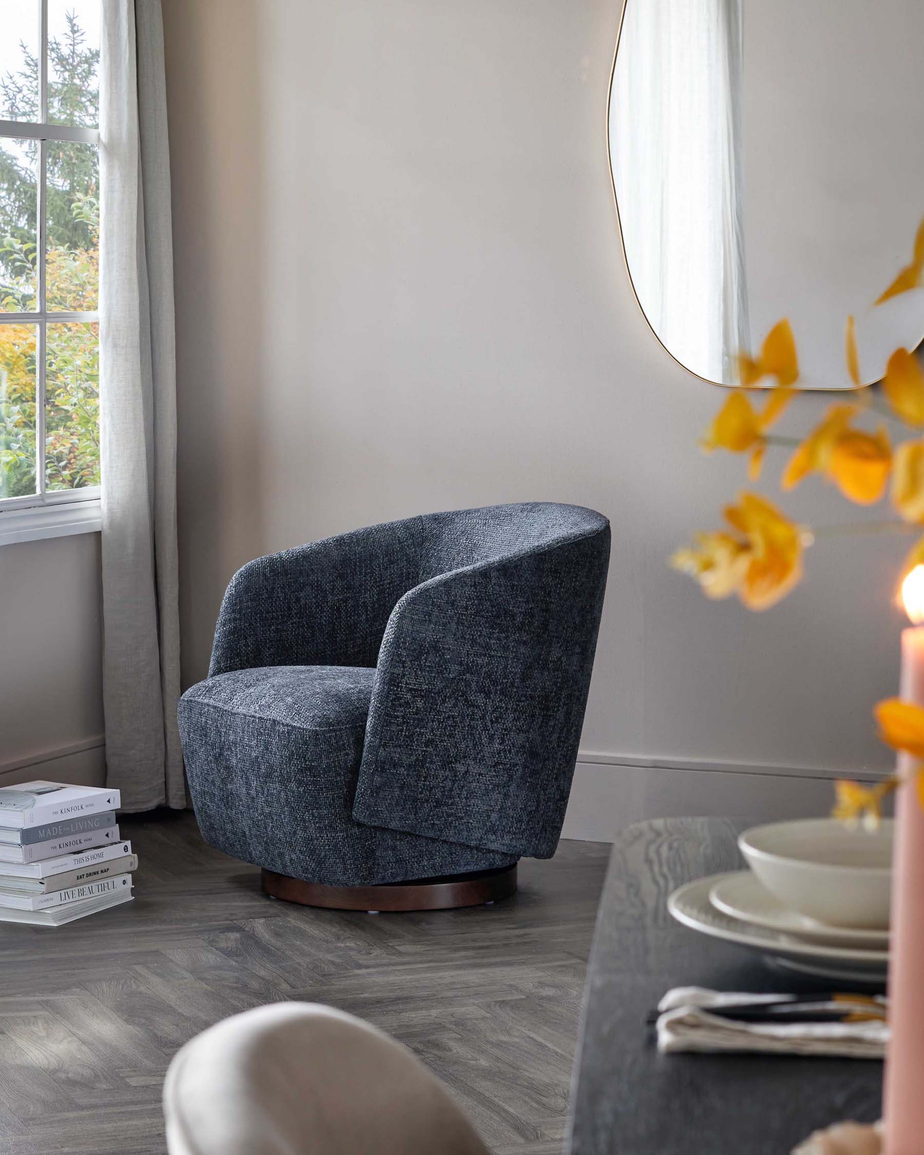 Modern, curved armchair in dark grey textured fabric with a smooth swivel base made of dark wood. The chair is positioned near a window with a view of greenery, accompanied by a stack of books on the floor, and is part of a serene room decor complemented by neutral tones.