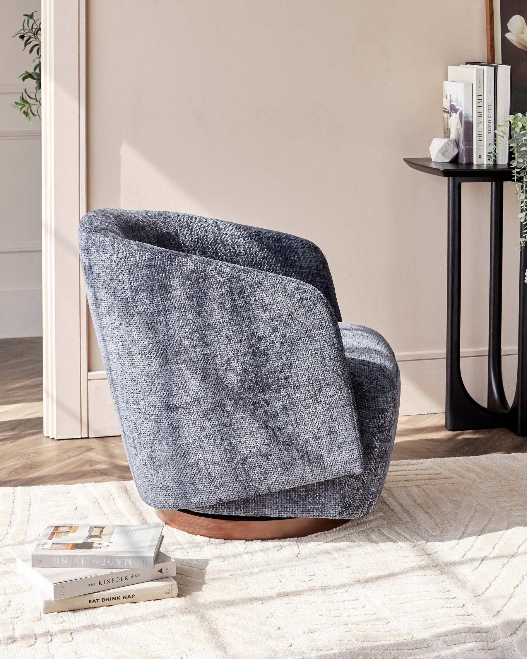 A modern accent chair in textured dark grey upholstery with a curved backrest and a circular wooden base, placed on a cream textured area rug, alongside a slim black side table with books.