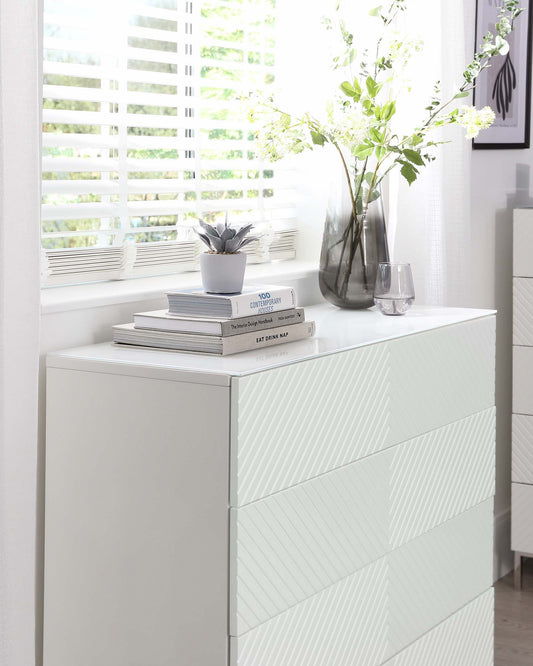 Modern white dresser with textured drawers, topped with books, a small potted plant, and a decorative vase of flowers.