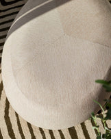 Close-up of a modern, rounded, beige fabric ottoman with subtle stitching detail, placed on a cream and tan striped area rug.