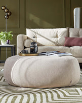 Contemporary living room setting with a large, plush, beige fabric sofa featuring multiple soft cushions. In front of the sofa is an oval-shaped fabric ottoman in a matching beige tone, situated on a patterned area rug with organic, wave-like designs in shades of green and beige. To the left is a small round side table holding a plant and a variety of decorative items.