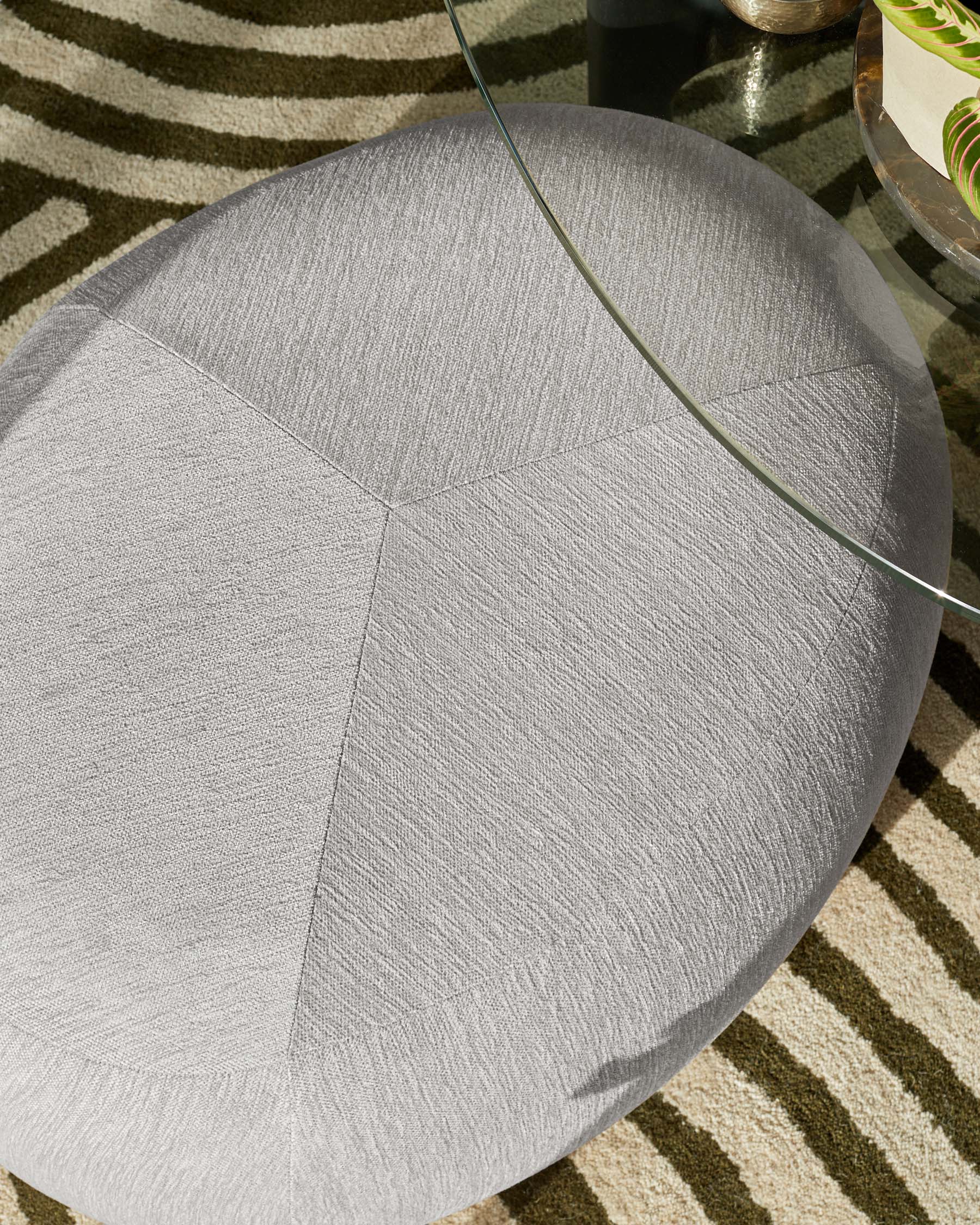 Round upholstered light grey ottoman with a geometric panelled design, partially visible glass side table with a thick glass top, on a beige and brown striped area rug.