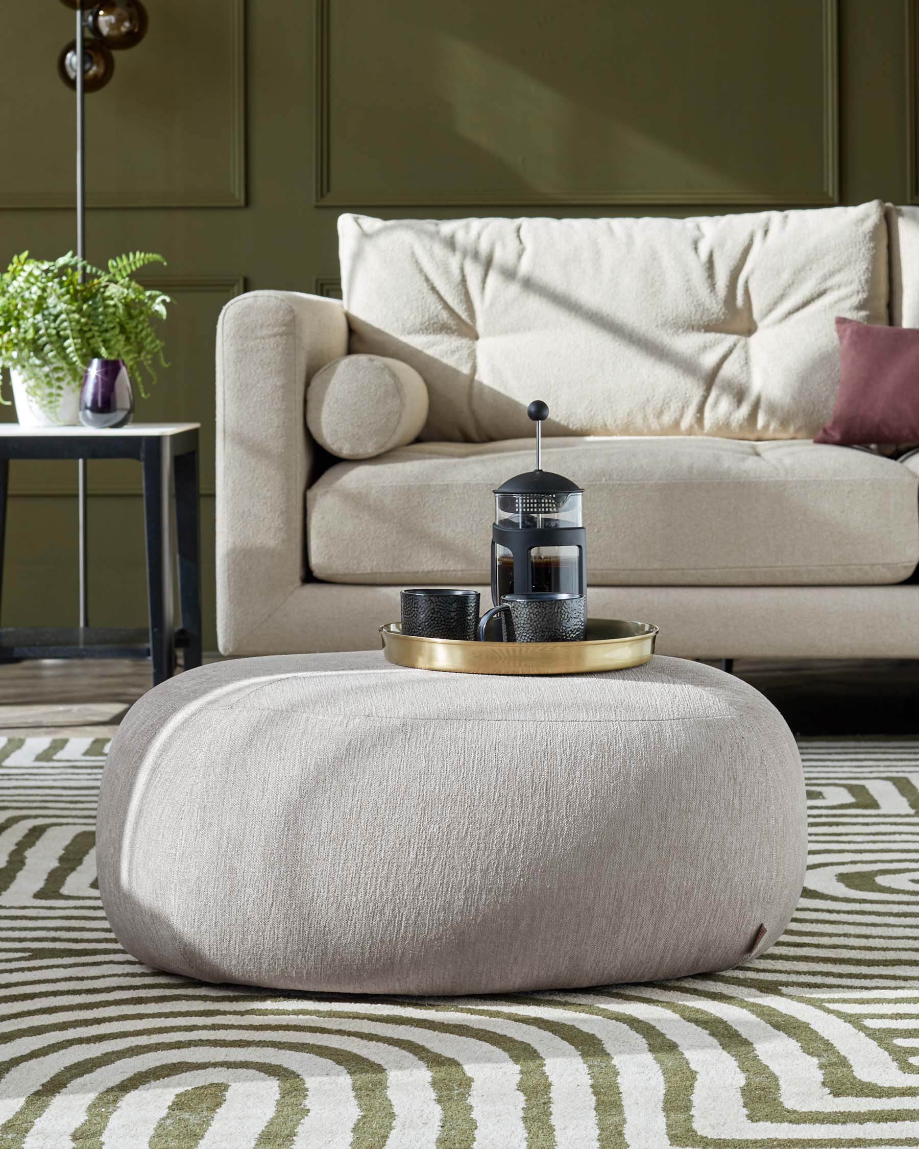 A contemporary living room setting with a plush beige three-seat sofa adorned with various pillows, complemented by a round light beige fabric ottoman in the foreground, and a small round side table with a potted fern to the left. The ensemble is arranged on an abstract-patterned area rug in shades of green and beige.