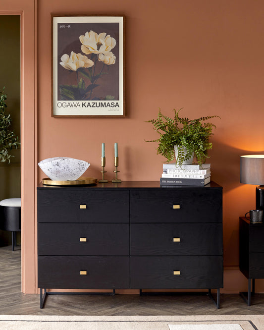 A stylish black dresser with gold accents, topped with decor including a bowl, candles, books, and a potted fern.