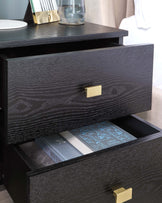 A sleek black nightstand with two drawers, featuring gold handles and a simple decorative setup with a glass and photo frame.