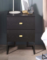 Modern black nightstand with two drawers, gold handles, a white lamp, a glass tumbler, and a decorative framed print.