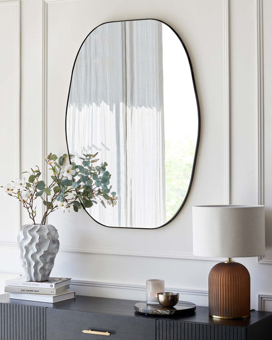 A modern, black textured sideboard with gold handles under a large, minimalist oval mirror. The sideboard is adorned with stylish decor including a sculptural grey vase with eucalyptus branches on a stack of coffee table books, an elegant white lamp with a brown base, and a small tray with a candle and a cup.