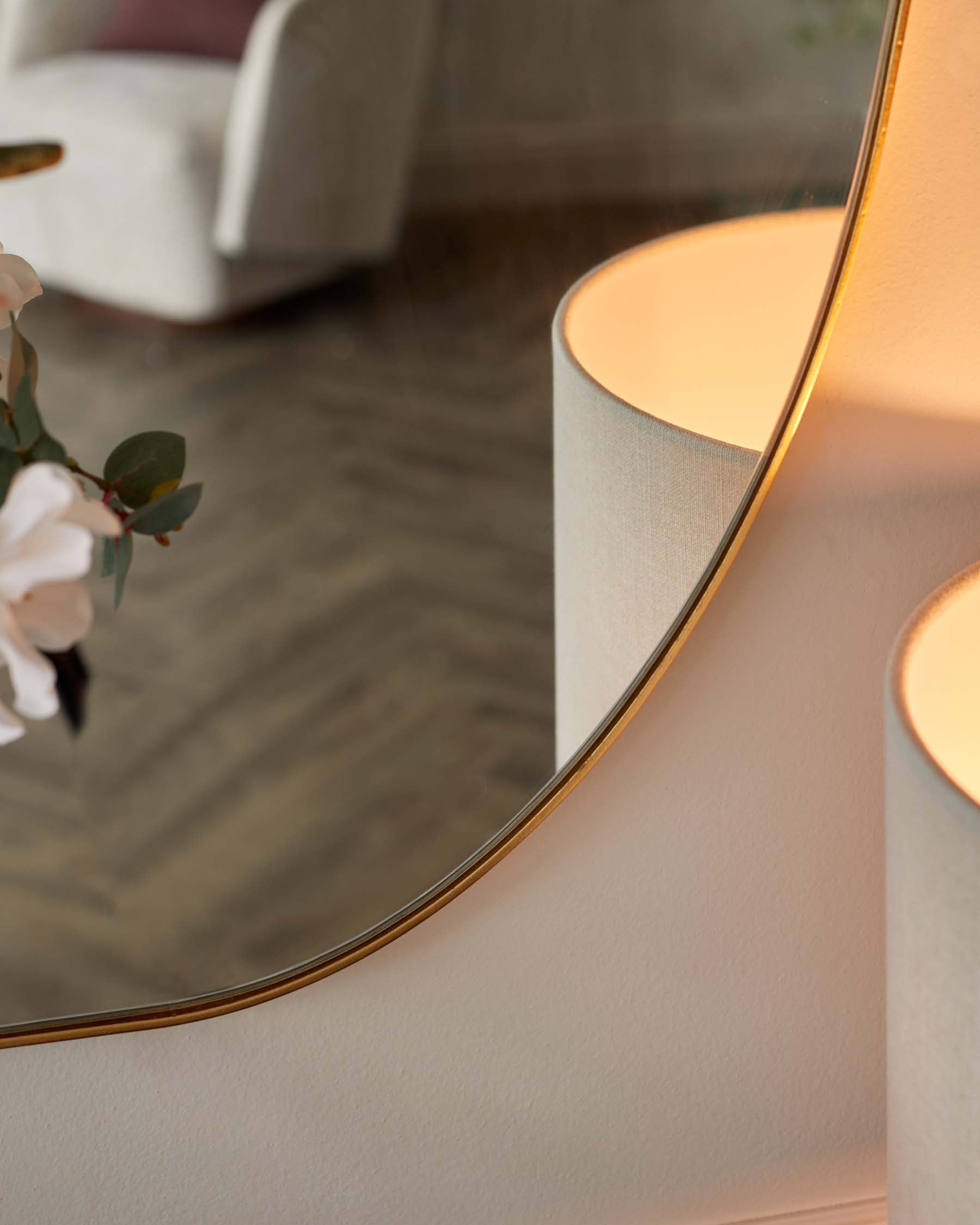 Elegant modern armchair with a curved silhouette and light grey upholstery, partially visible in a chic interior setting, with a touch of sophistication added by the reflection in a circular mirror with a slim, gold frame.