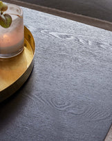 Elegant dark wood grain textured table surface with a subtle sheen, showcasing natural wood patterns and a sophisticated modern aesthetic. A gold-toned metallic tray rests on the table, holding a glass with a frosted beverage, accentuating the table's luxurious appeal.