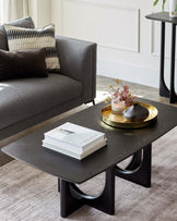 A contemporary living room setup featuring a dark wooden coffee table with a unique curved leg design on a light grey area rug. The table is accessorized with a golden tray, candles, a vase with flowers, and topped with a stack of books. Behind it is a portion of a grey upholstered couch with plush pillows, including a striped one, facing a plain white wall.