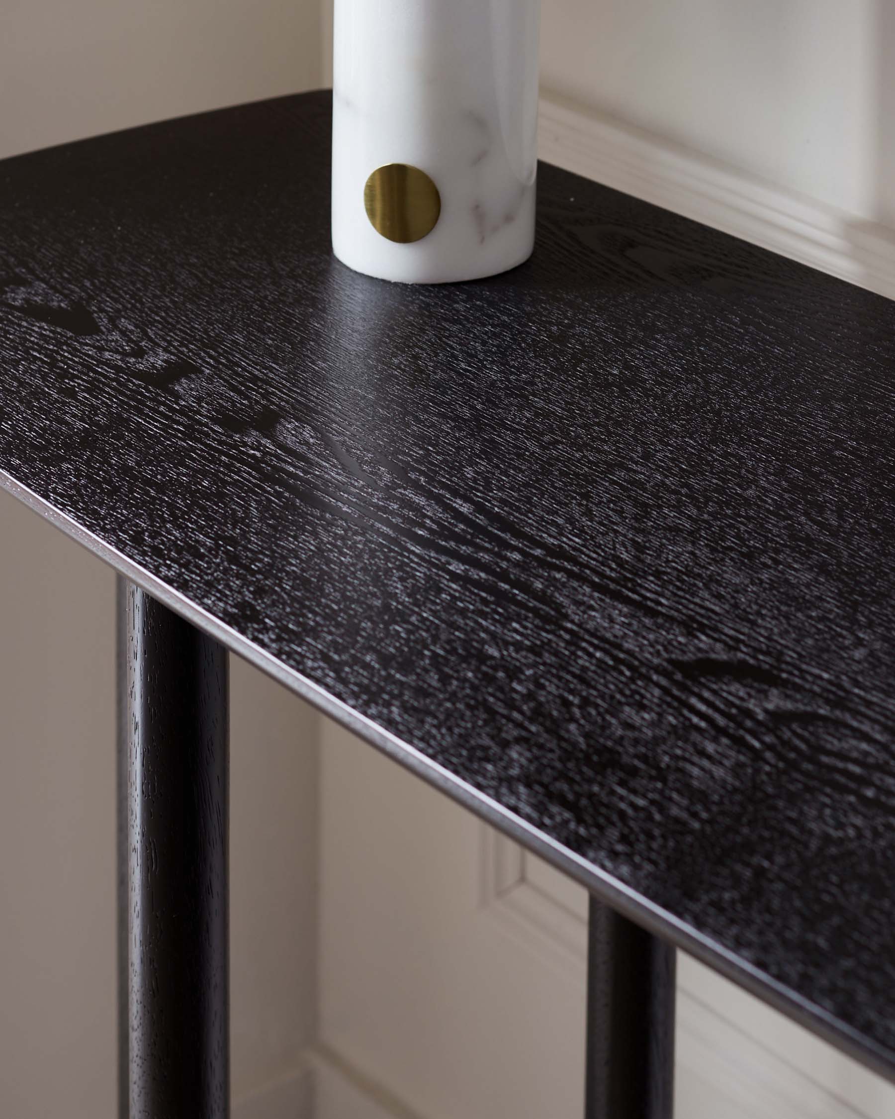 A modern console table with a dark wood grain finish and sleek, black metal legs, featuring a white marble cylinder with a brass accent on top.