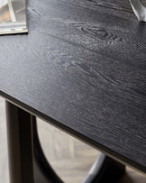 Close-up of a dark wood dining table with a textured surface and a subtle sheen. The table features an elegant, understated design with curved legs, suggesting a modern yet timeless style. Part of a magazine is visible in the corner, indicating the table's use in a contemporary living space.