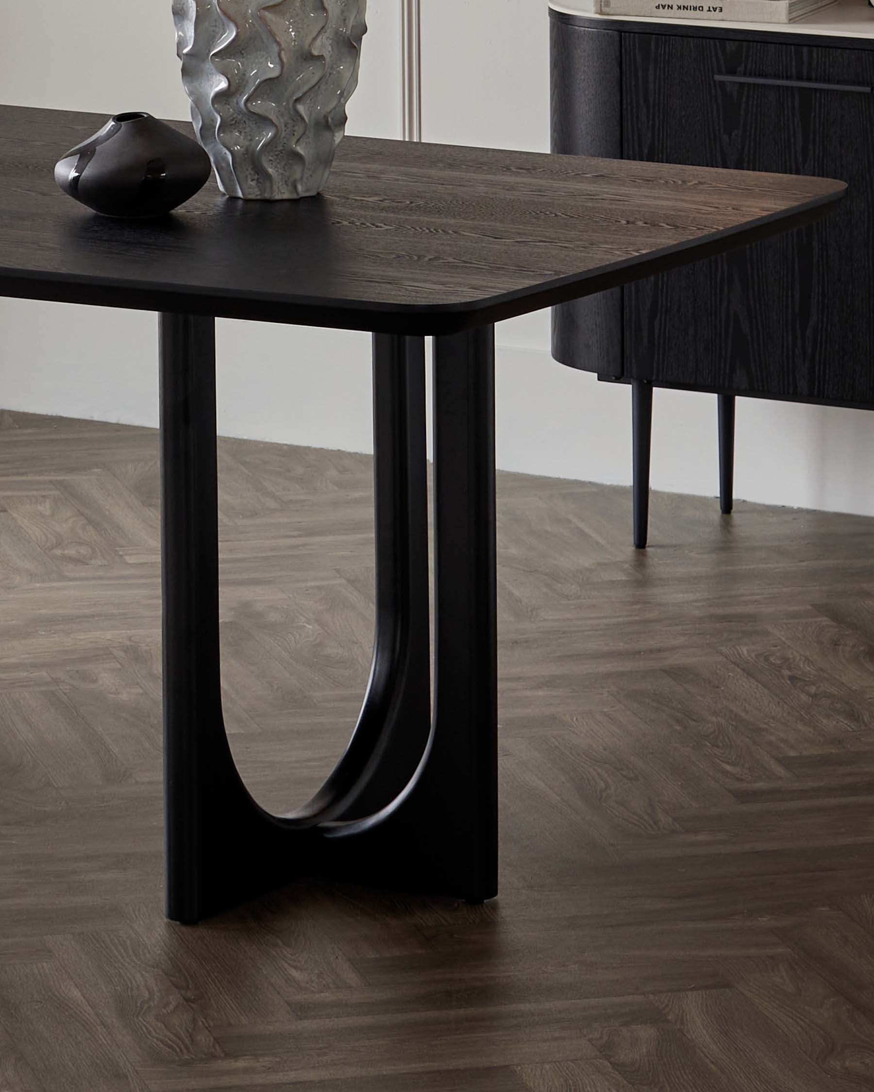 Elegant modern dining table with a textured dark wood finish and curved black metal legs, set on a hardwood floor with decorative vases as accents. A sideboard with a complementary dark wood veneer is partially visible in the background.