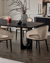 A contemporary dining room set featuring a dark wooden table with a subtle grain pattern and sleek black metal legs. Surrounding the table are plush velvet-upholstered chairs in a light taupe colour, with rounded backrests and black legs that complement the table. The furniture is set on a dark herringbone wood floor, creating an elegant and modern aesthetic.