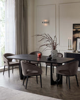 A modern dining room set featuring a rectangular, dark wood table with an organic edge and robust black legs. Accompanied by five velvety, upholstered dining chairs with curved backrests and black tapered legs. The table is decorated with a tall ceramic vase with dried branches, a lit red candle, and a sleek black oval centrepiece.