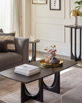 Contemporary living room furniture including a rectangular coffee table with black legs and a dark wood grain top, accessorized with books and a golden tray holding a candle and vase. Behind it, a round side table with a dark top and black metal legs is adorned with a glass and candleholder.