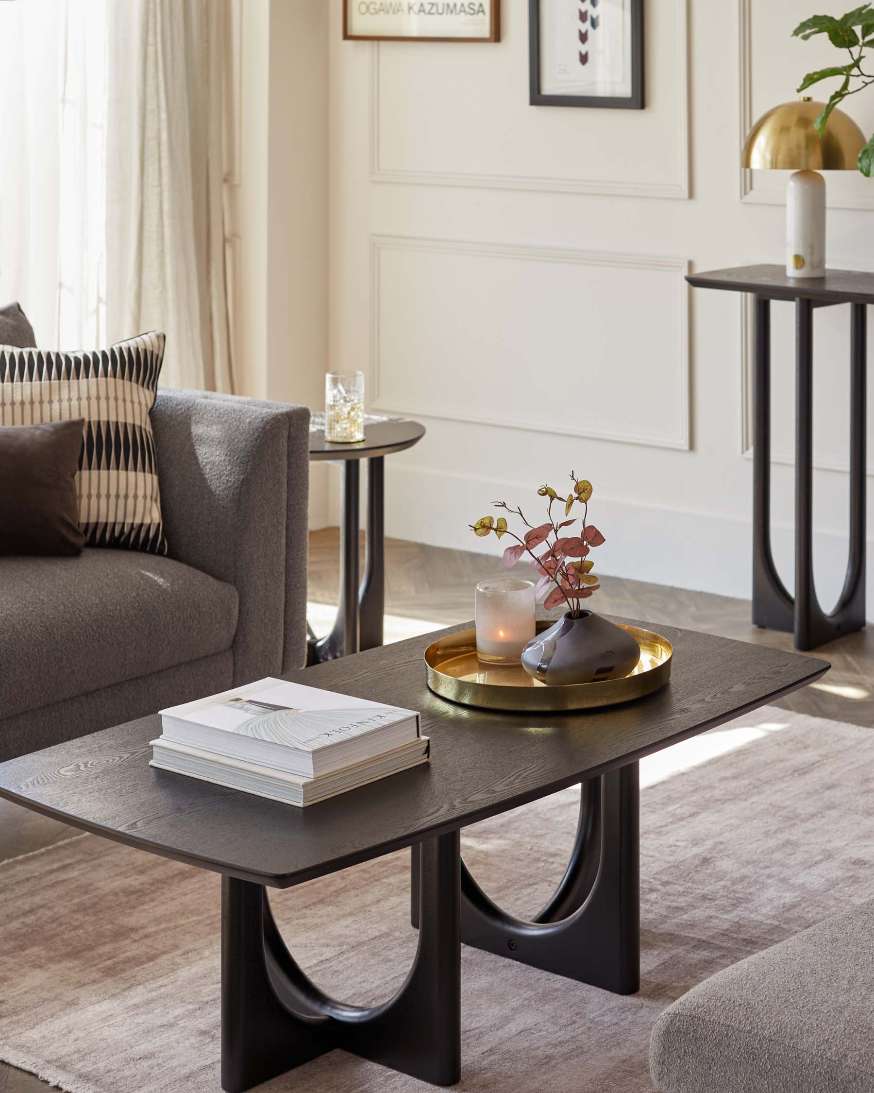 Contemporary living room furniture including a rectangular coffee table with black legs and a dark wood grain top, accessorized with books and a golden tray holding a candle and vase. Behind it, a round side table with a dark top and black metal legs is adorned with a glass and candleholder.