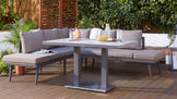 Modern outdoor seating with gray cushions and a square dining table, surrounded by greenery on a wooden deck.