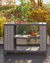 Outdoor kitchen island with a built-in sink and modern faucet, presented in a grey wood finish, featuring open shelving, a towel rack on one side, and placed against a lush garden background.