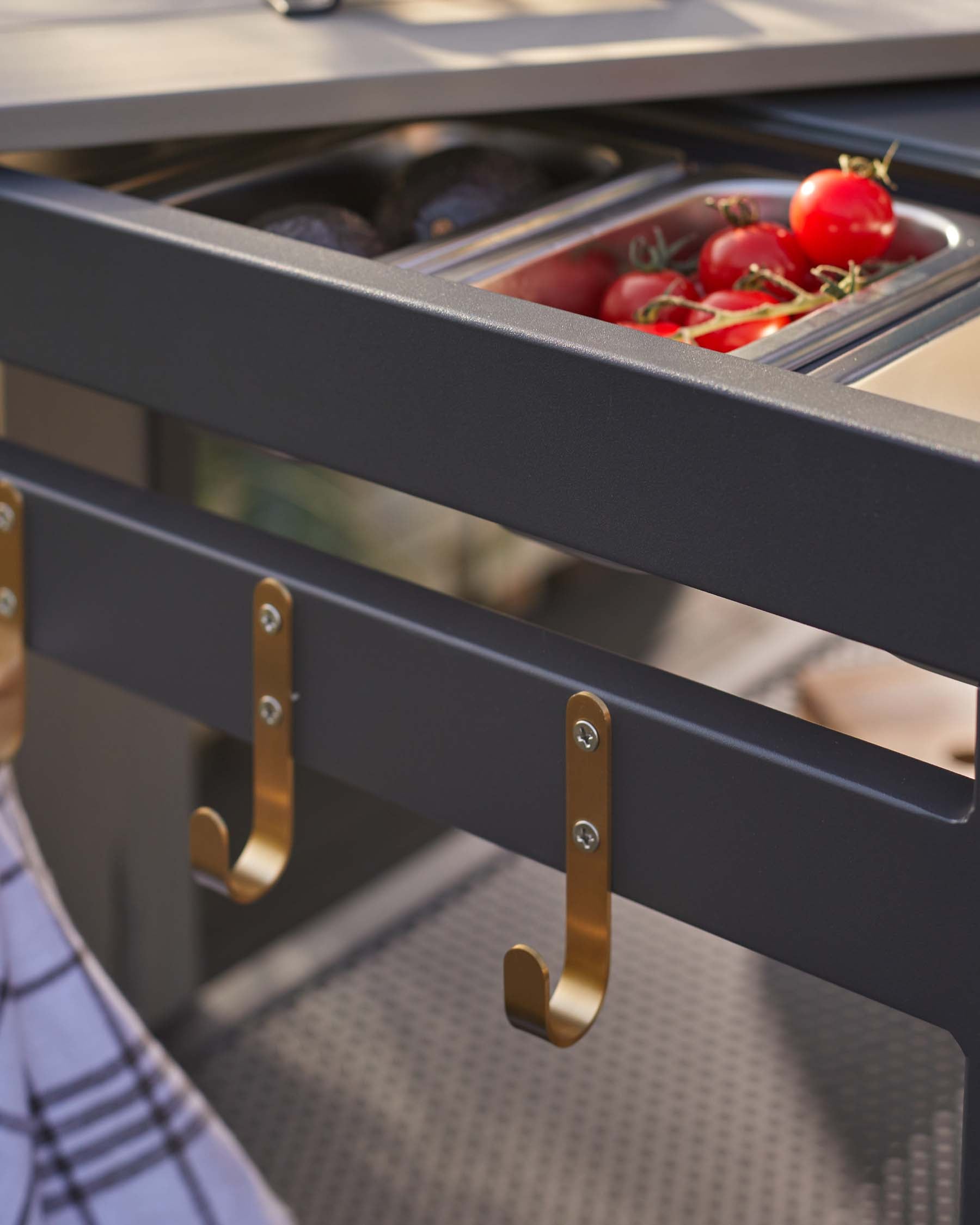 Modern outdoor kitchen module featuring a sleek, dark grey design with stainless steel countertop and integrated gas burners. Underneath, a spacious drawer with a clear-view insert reveals neatly organized produce, flanked by elegant brass-finished handles.