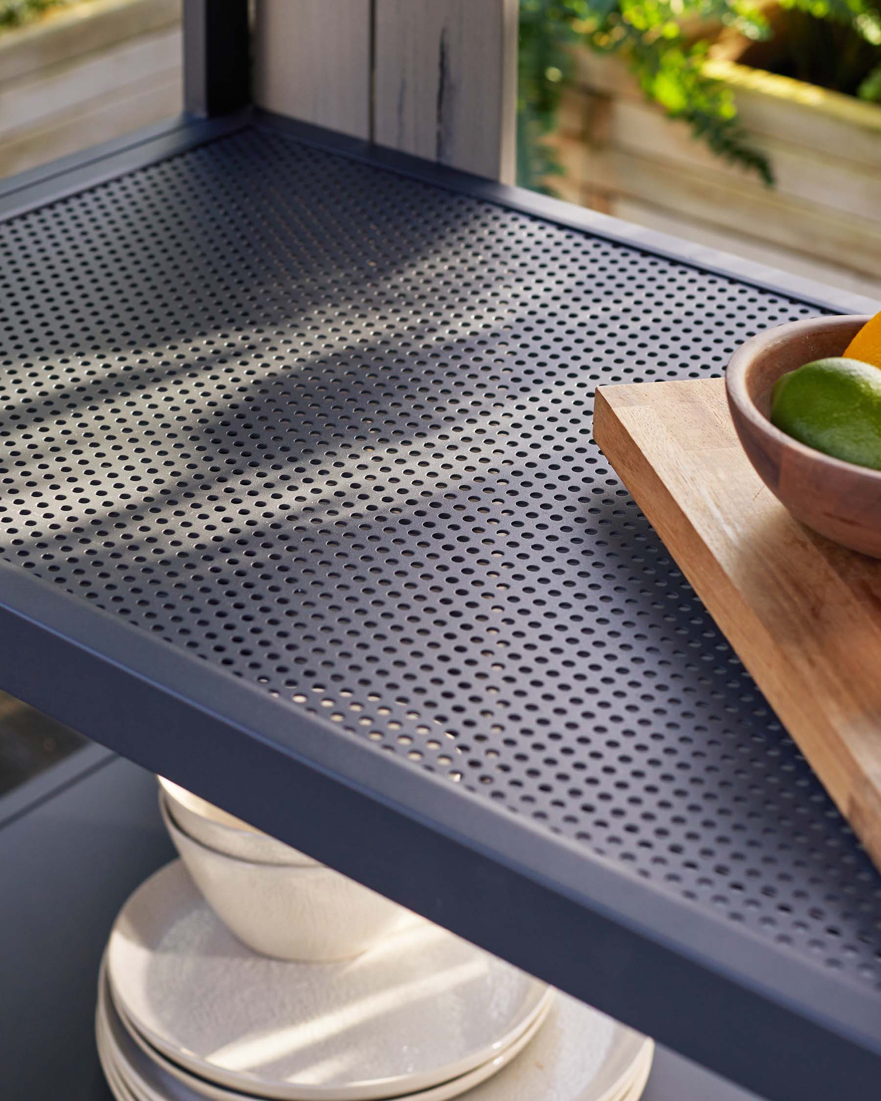 Modern metal trolley with perforated shelves in dark grey, featuring a wooden cutting board with a bowl of limes on the top tier, and stacked ceramic plates on the bottom shelf, set against a sunny window with a view of green foliage.