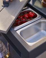 Modern outdoor kitchen module featuring a sleek grey countertop with an integrated stainless steel container holding fresh tomatoes and an empty compartment beside it, set in a sturdy black frame with a handy brass-finished towel holder.