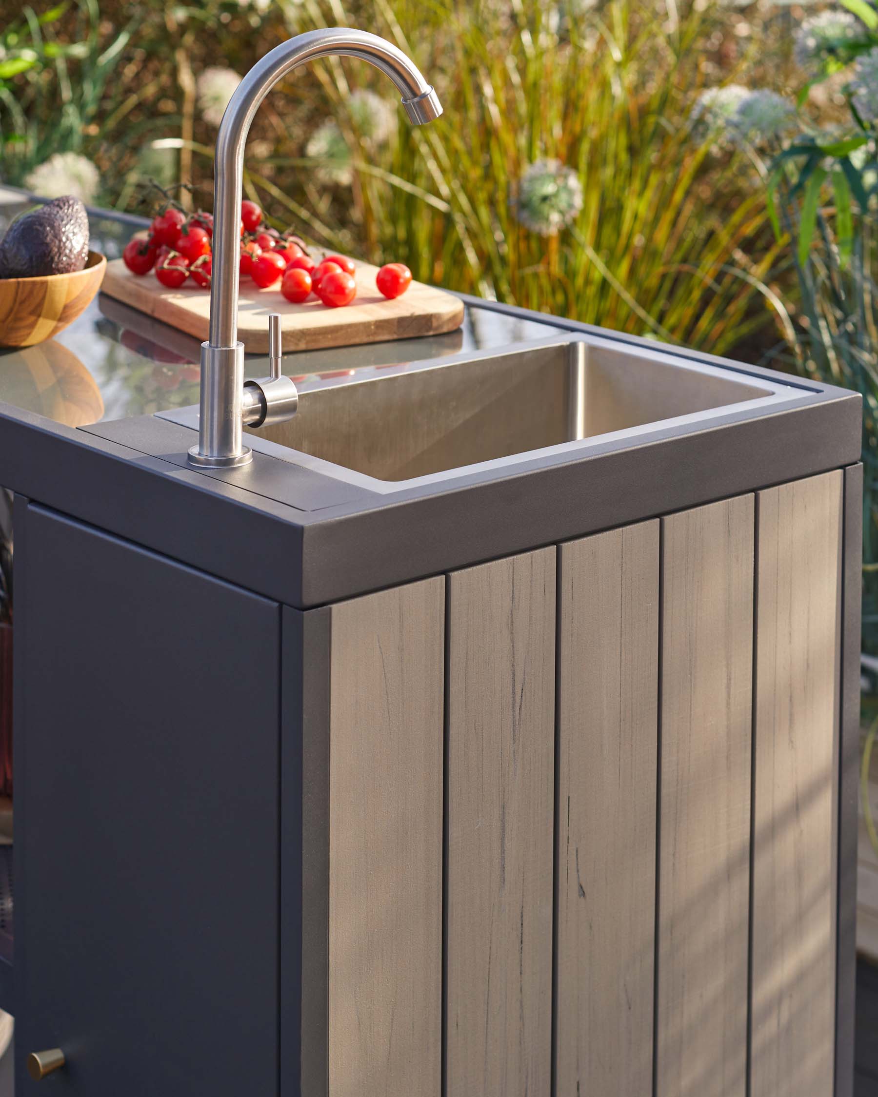 Modern outdoor kitchen unit featuring a charcoal grey base and stainless steel sink with a sleek, curved faucet. The wooden front panels have a washed-out finish, and there's a wooden countertop extension displaying fresh produce.