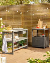 A stylish outdoor kitchen cart with shelves and a drawer, paired with a compact, dark storage cabinet featuring gold handles.