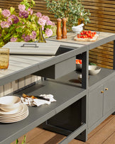 Modern outdoor kitchen furniture with a grey finish, featuring storage cabinets, countertop, plates, utensils, and fresh produce.