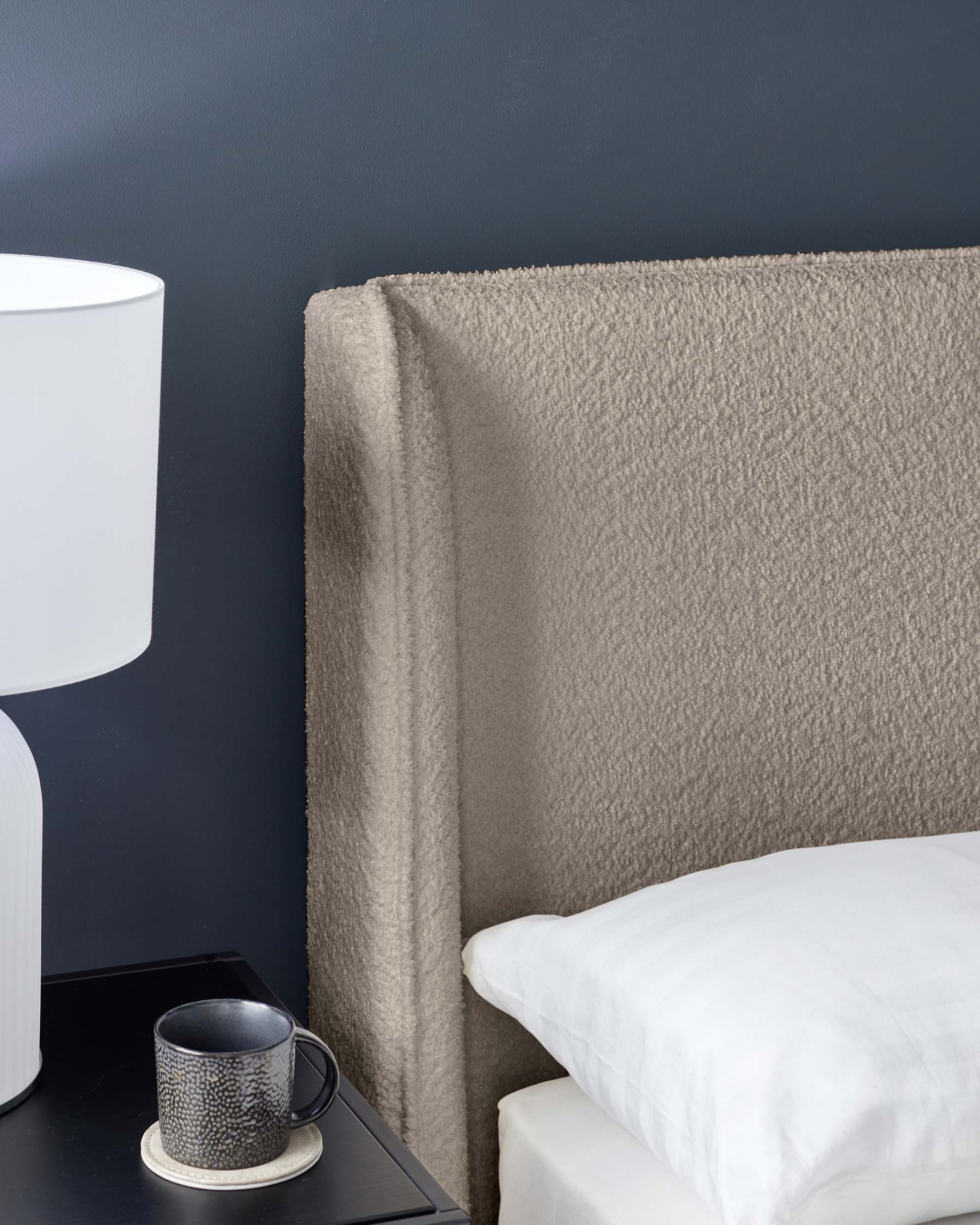 Modern bedroom featuring a plush beige upholstered headboard, a sleek black bedside table with a textured ceramic cup and saucer on top, and a white contemporary lamp with a rounded base and cylindrical shade.