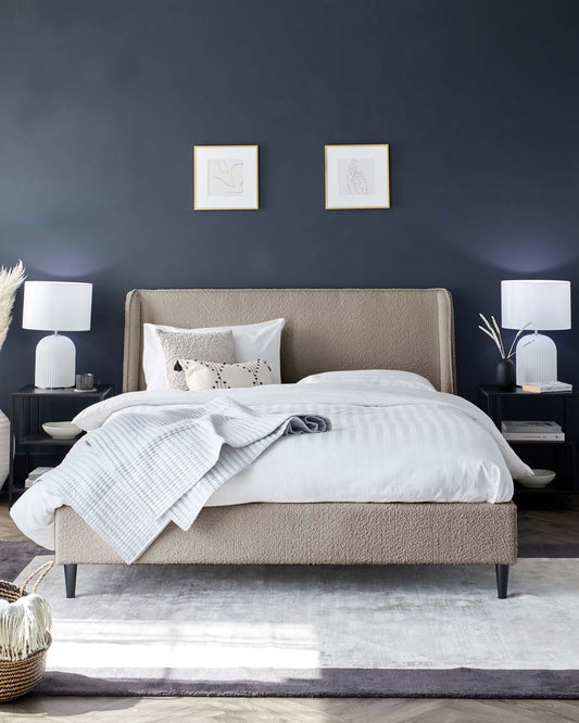 Modern bedroom featuring an upholstered platform bed in a neutral beige fabric with a high, cushioned headboard and dark tapered legs. Coordinating bedside tables in black with elegant white table lamps, and a soft grey floor rug complete the contemporary aesthetic.