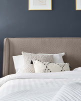 Contemporary upholstered headboard in a textured beige fabric with a slightly curved top edge, accompanied by a bed dressed in a white textured duvet cover and an arrangement of decorative pillows, set against a dark blue wall with two framed art pieces above.